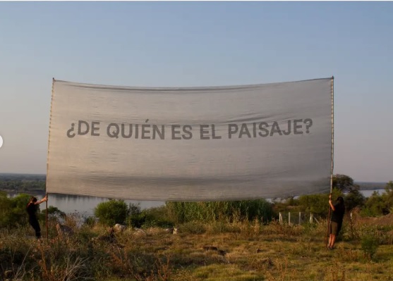 Dos personas sostienen una pancarta de grandes dimensiones frente a la vera del rio en un parque. La pancarta dice ¿De quien es el paisaje?