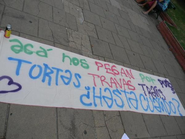 Fotografía de la bandera pintada en el marco de esta manifestación. Consigna: acá nos pegan por putos, tortas, travas, trasn ¿hasta cuándo?